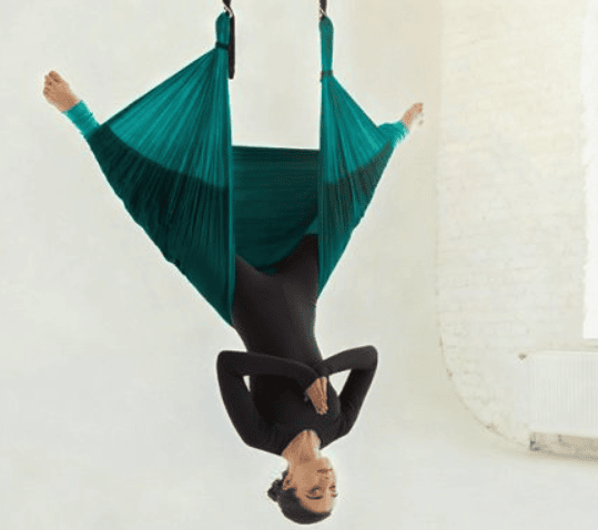 A woman practicing aerial yoga, suspended upside down in a teal fabric hammock. She is wearing a black long sleeved bodysuit and has her hands in a prayer position near her chest. Her legs are spread wide within the silk, and the background is a bright, minimalist white studio with a brick wall.
