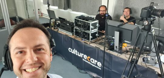 Foto von Udo Nauber, der im Vordergrund steht und eine Headset trägt. Schräg hinter ihm ist diverse Veranstaltungstechnik auf einem Tisch mit einem Banner, das den Namen seiner Firma culturecare trägt, aufgebaut, unter anderem eine Kamera, Mischpulte und Computer. Hinter dem Tisch sitzen nicht namentlich bekannte Mitarbeiter der Firma. Beide tragen ebenso Headsets, und einer zeigt einen Daumen nach oben.
