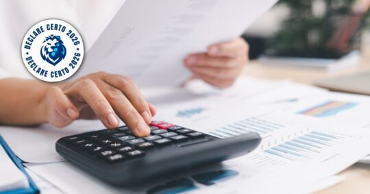 Pessoa usa calculadora sobre documentos financeiros. No canto, selo azul "Declare Certo 2026" com desenho de leão.