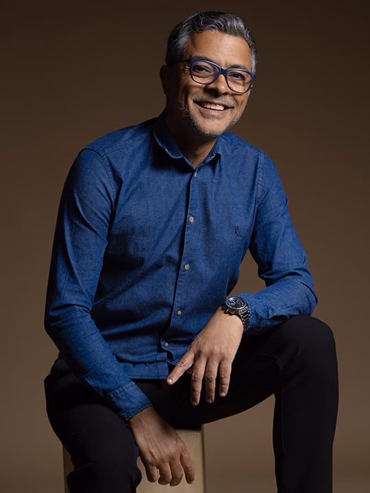 Alessandro Bastos, o Alê, fotógrafo corporativo, de camisa azul e óculos, sorrindo no estúdio Meu Novo Retrato em Belo Horizonte