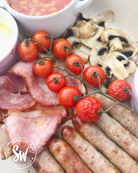 Close up of oven roasted tomatoes on top of cooked sausages, bacon and mushrooms in the full english breakfast traybake