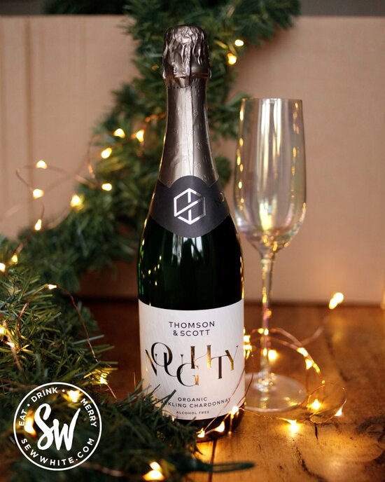 Bottle of Noughty Organic alcohol-free sparkling wine in the drink gift guide list next to a garland of green with fairy lights and champagne glass. 