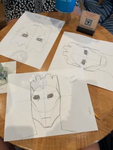 Drawings of superhero masks on paper sketches, including Groot, Iron Man, and others, at a restaurant table.