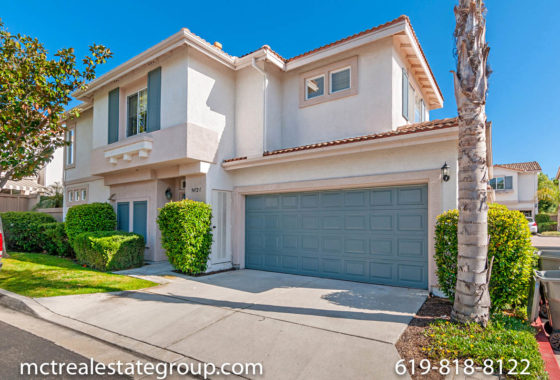 Two-story suburban home at 9472 Compass Dr-S featuring light beige stucco siding, a large blue-gray garage door, decorative blue shutters, and manicured hedges on a sunny day.