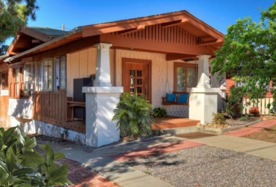 Exterior side view of the historic Craftsman bungalow at 1930 Granada Avenue in South Park with a covered front porch, white pedestal columns, and a wooden porch swing under a clear blue sky.