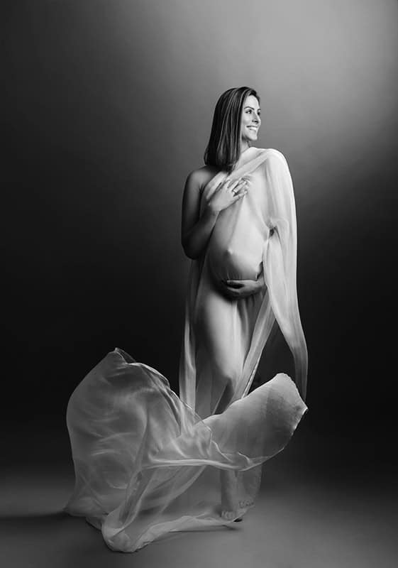 Kvinne gravidfotografering i studio, svart-hvitt bilde med elegant graviditetsportrett, myk belysning og delikat kjole som omkranser den gravide magen. Perfekt for gravidfoto og mesterlige morsbilder.
