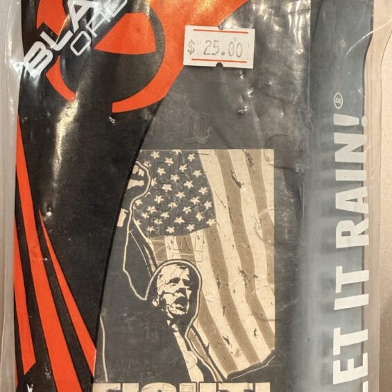 Black graphic T-shirt in plastic packaging with a red and black design, an American flag motif, and bold white 'FIGHT' text; also shows 'MADE IN USA' text on the bag.