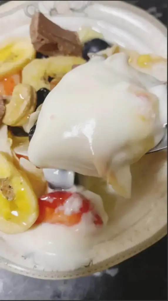 Spoon scooping yogurt with strawberries, blueberries, nuts, and chocolate from a parfait bowl.