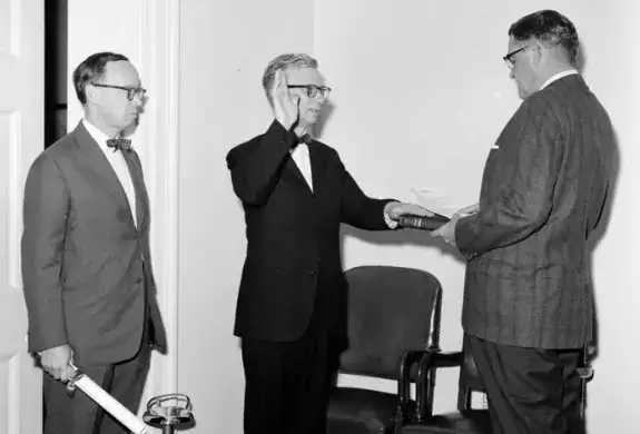 A man in glasses places his left hand on the Bible and his right in the air. Two men in suits stand in front and behind him.