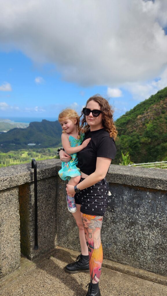 Phoenyx holding River while standing at the Pali Lookout in Oahu, Hawaii, with a scenic backdrop of lush green mountains and the blue ocean under a partly cloudy sky.