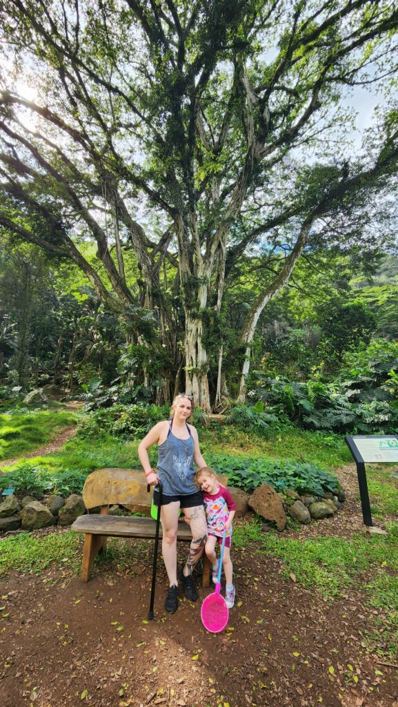 Phoenyx and River standing together on the Waimea Falls Trail in Oahu. They are surrounded by lush green foliage and a towering tree with sprawling branches, capturing the natural beauty of the Hawaiian landscape.