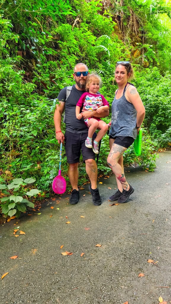 A family of three stands on a shaded trail in Waimea Valley, surrounded by lush jungle plants, with the dad holding a pink bug net and the daughter in his arms.