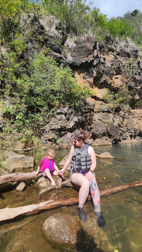 A mother in water shoes and a daughter in a pink life vest sit on rocks near a shallow stream at Waimea Falls, holding hands and relaxing together.