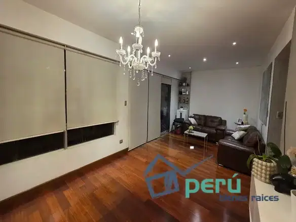 Bright living room with dark wood floor, a crystal chandelier, and large white blinds along one wall; modern sliding doors and a brown leather sectional seating area in the corner.