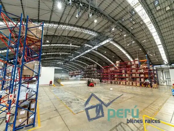Wide warehouse interior with tall red-and-blue shelving racks filled with boxes and a small forklift in the center, bright overhead lights