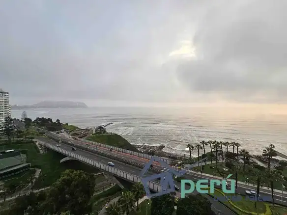 Aerial view of a coastal Peruvian city with a bridge over a highway, palm trees along the promenade, and the Pacific Ocean at sunset.