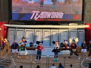 Colorful Disney characters Mickey Mouse, Minnie Mouse, Donald Duck, Daisy Duck, Pluto, and Goofy perform on stage at a cruise ship entertainment show.