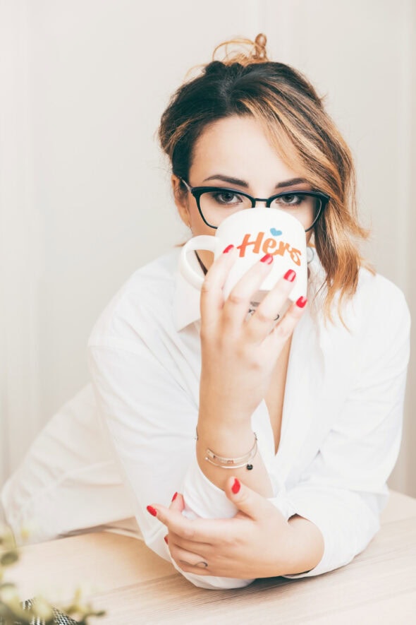 Donna in camicia bianca che tiene una tazza con la scritta "Happy" mentre guarda in camera, con occhiali e un'acconciatura ondulata.