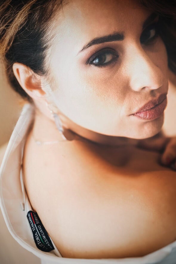 Ritratto di una donna con trucco elegante e orecchini, sguardo intenso e capelli raccolti. La luce mette in risalto la sua pelle e i dettagli del vestito.