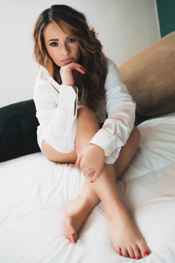 Modella seduta su un letto, indossando una camicia bianca, con i capelli mossi e uno sguardo pensieroso. La scena trasmette un'atmosfera di intimità e relax.