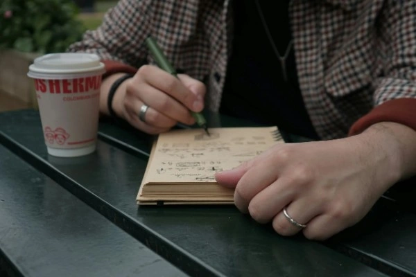 Person in plaid shirt writing thoughtfully in small notebook with coffee cup nearby at café table, representing the journaling and emotional preparation process for managing travel anxiety