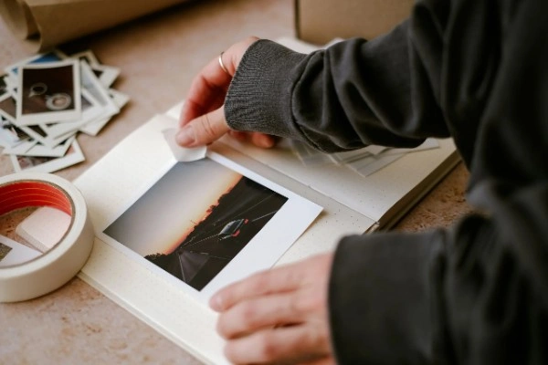Person thoughtfully organizing travel photos in album at home with scattered instant photos nearby, representing post-travel reflection and integration of experiences
