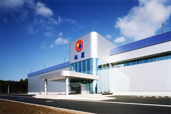 松屋富士山工場でガラスの入り口があるモダンな建物。正面にはカラフルな円形のロゴと松屋の文字が描かれ、上には澄んだ青空が広がっています。