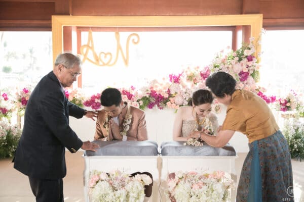Elders place the mongkol headpieces on the kneeling bride and groom to begin the Rod Nam Sang water blessing at Dusit Thani Hua Hin