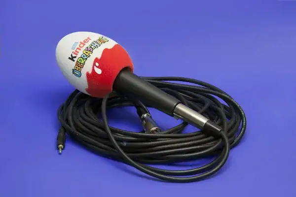 Handheld microphone with a custom printed foam windscreen (logo and colours) on a blue background, shown with a coiled cable for broadcast and interview use.