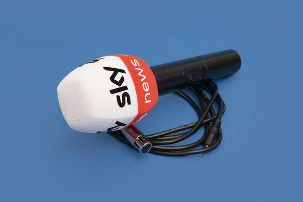 Handheld microphone with a white foam windscreen and an orange band featuring “news” text, shown with a cable on a blue background.