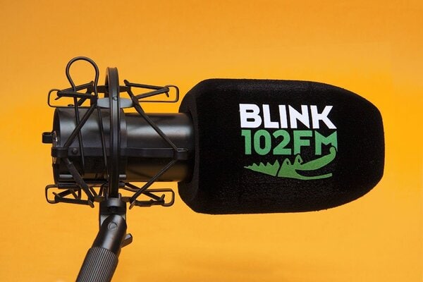Black foam microphone windscreen with “BLINK 102FM” logo on a studio microphone in a shock mount, shown on an orange background.