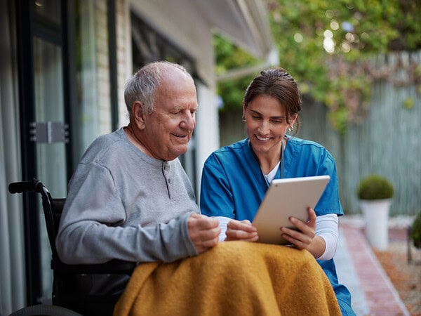 Freundliche Pflegekraft als Alltagsbegleitung für Senioren bei AlternativezumHeim Freundliche Krankenschwester und älterer Mann im Rollstuhl, der ein digitales Tablet benutzt