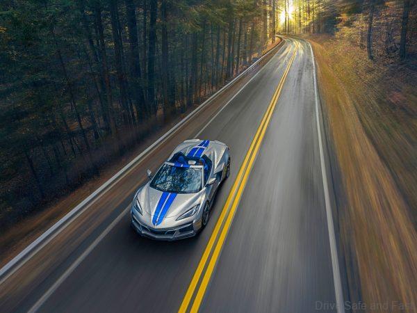Overhead front view of 2024 Chevrolet Corvette E-Ray 3LZ convertible in Silver Flare with Electric Blue stripe package driving on a road between trees. Pre-production model shown. Actual production model may vary. Model year 2024 Corvette E-Ray available 2023.