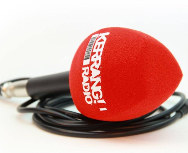 Handheld microphone with a red foam windscreen printed with the vertical white “KERRANG! RADIO” logo, lying on a white surface with a coiled cable.