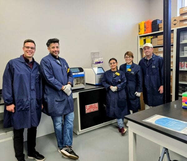 A group of five scientists, all wearing blue lab coats, are standing together in a lab. In the center of the group are two female scientists, and on either side are three male scientists. A large scientific machine is on the counter behind them.