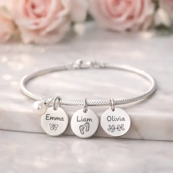 Bracelet de naissance en argent avec charms gravés posé délicatement sur une surface en marbre blanc avec des fleurs fraîches en arrière-plan flou.