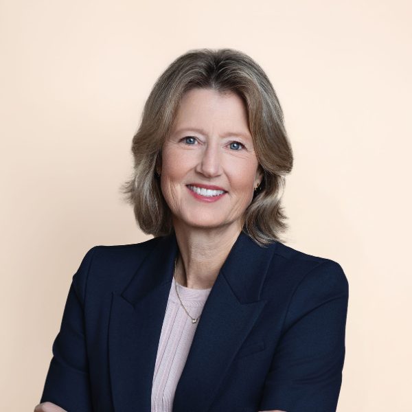 A woman with shoulder-length blonde hair wearing a dark blazer and light top stands against a plain beige background, smiling with arms crossed.