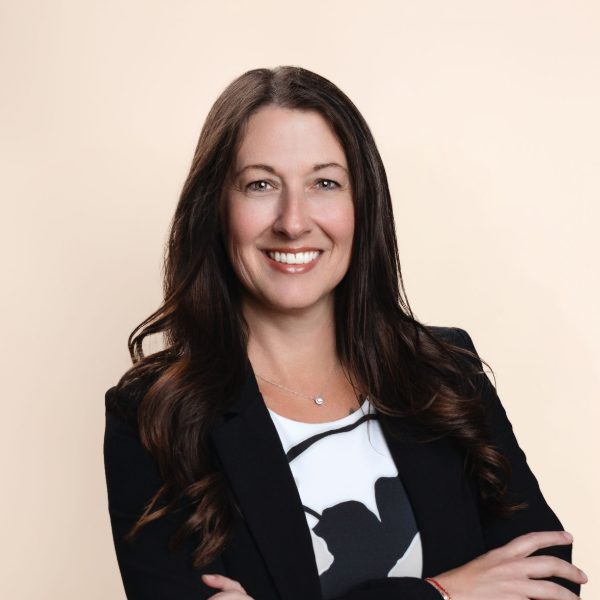 Woman with long brown hair in a black blazer and patterned top stands with arms crossed, smiling, against a plain light background.