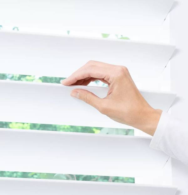 A hand opening white window blinds for sunlight and fresh air.