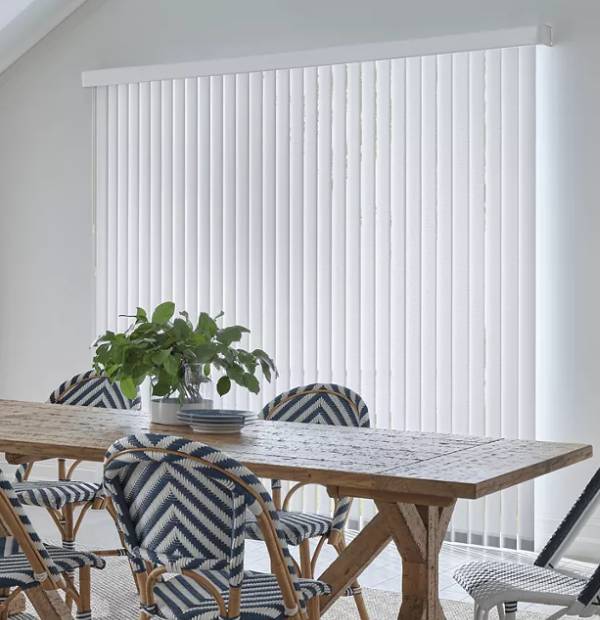 Vertical blinds covering large window in bright dining room with wooden table.