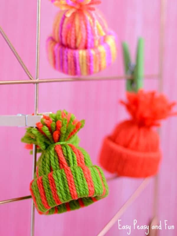 mini hat ornaments for Christmas made with yarn hanging in a pink background