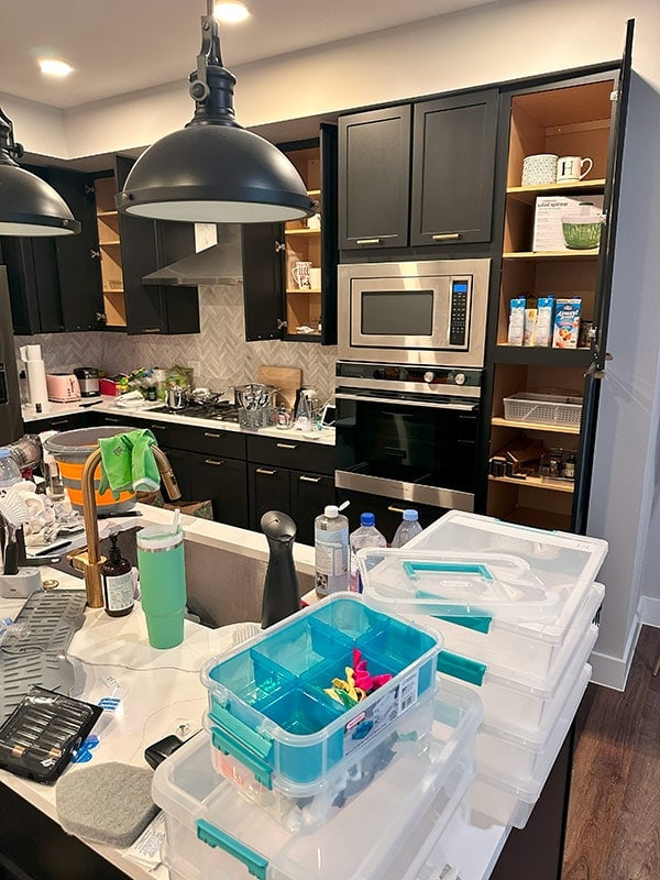 Cluttered kitchen with black cabinetry, built-in microwave, oven, and open shelves filled with various items and snacks, all under modern black pendant lights.