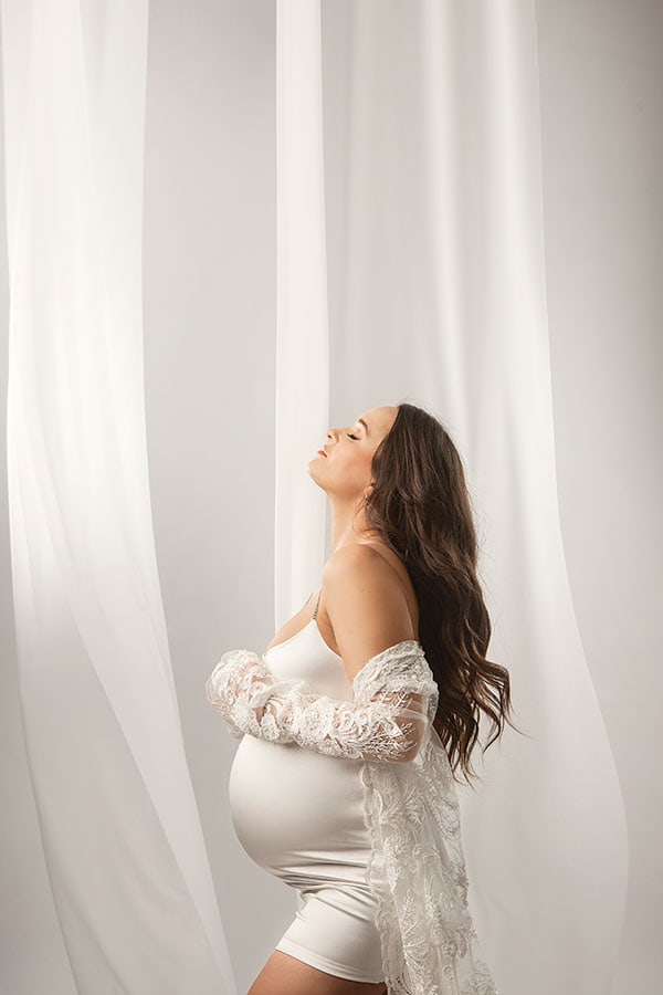 Fredelig gravid kvinne i kjole og blondestole foran hvite gardiner, capturing en betagende graviditetsfotografering.