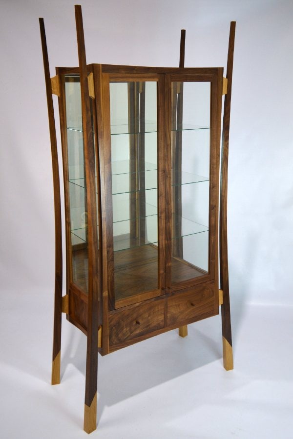 Custom made from American Black Walnut and finished in Tru oil to give a nice warm glow to the timber. This display cabinet with its modern bespoke design and heavy use of curves and angles was to create an eye-catching design. The legs are free-floating from the carcass at an angle of 15 degrees to create this unique design. The tapered curved legs that flare out at the bottom helps to give the cabinet better balance and stability. The use of white oak as an accent timber is used to create another design element in the design with the feet caps and joints between the carcass and legs. This cabinet has been constructed using hand-cut piston fit, houndstooth dovetail draws, and mortise and tenon joints in the frames. All the panels are book-matched solid walnut. A mirror at the back of the cabinet is to reflect light forward to better showcase the items the client collects. The draws in the bottom are to hold the paperwork and certificates of the collectibles so he would be able to keep everything in one place.