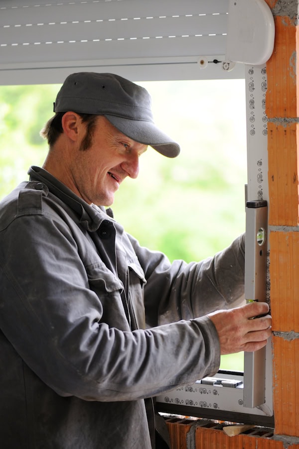 Home inspector in a grey cap and jacket using a spirit level to verify the alignment of a newly installed window frame in a brick wall.