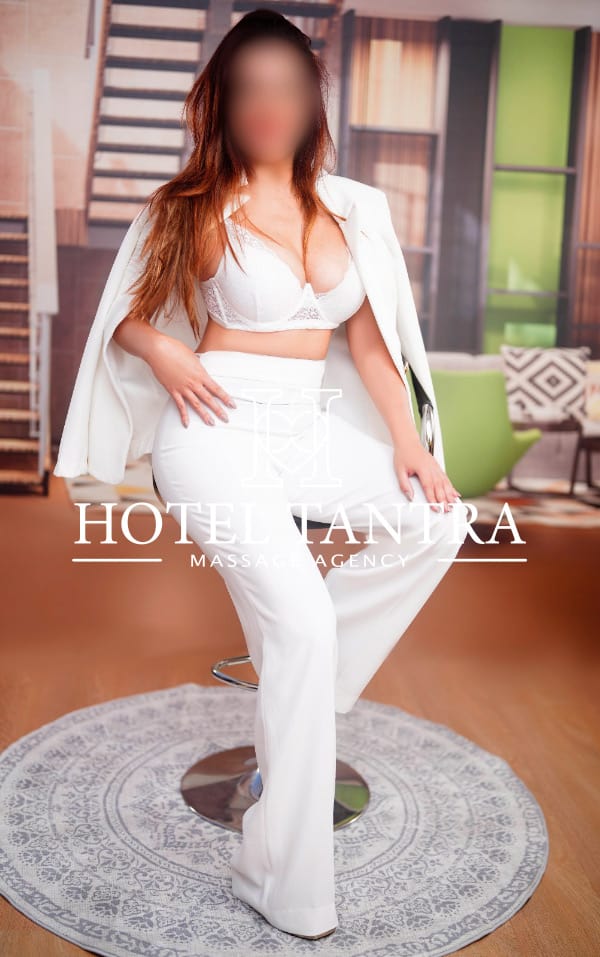 Woman in a white blazer and trousers with lingerie, seated on a bar stool in a modern lounge; watermark Hotel Tantra Massage Agency.