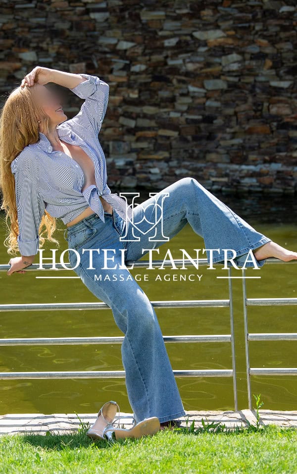 Person leaning on a railing beside a green-water body, wearing jeans and a striped shirt, with the Hotel Tantra Massage Agency logo overlayed on the image.