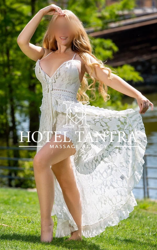 Woman in a white lace dress posing on grass outdoors, lifting the skirt with one hand; watermark 'Hotel Tantra' across the center.