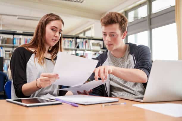 Two students collaborating in a library, reviewing documents and using a laptop, with bookshelves and study materials on the table