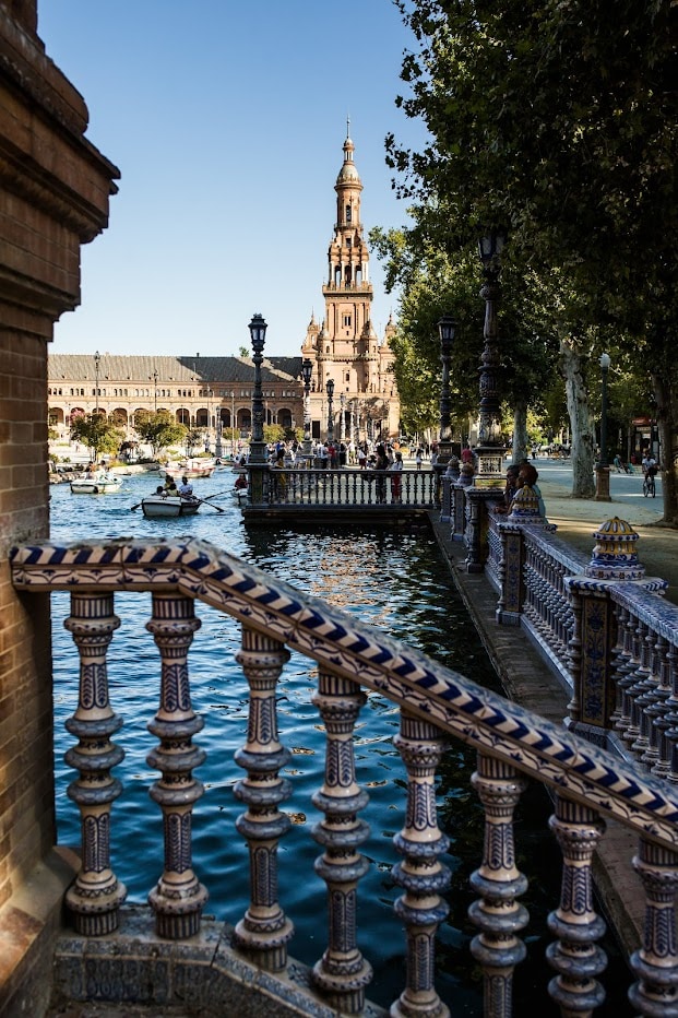 Blick durch ein kunstvoll verziertes Geländer auf den Kanal und den Turm der Plaza de España in Sevilla bei Tageslicht.